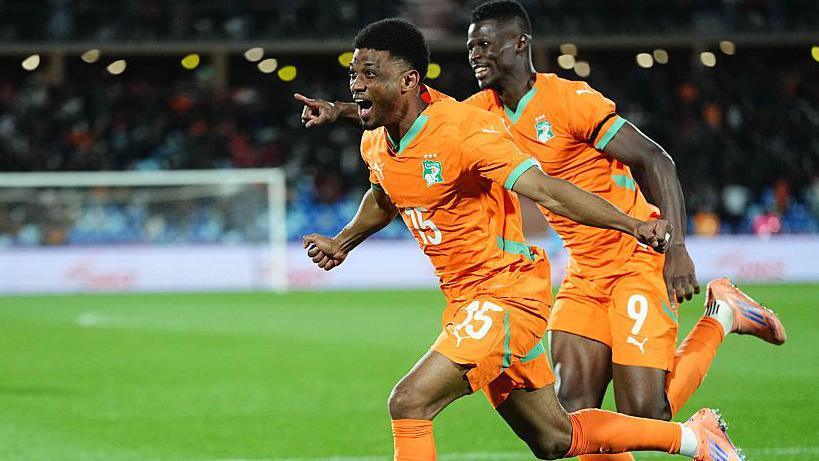 Footballer Amad Diallo, wearing an orange jersey with green trim and the number 15 in white on its front and orange shorts, spreads his arms wide to the side and runs to celebrate after scoring for Ivory Coast against Cameroon at Afcon 2025. Team-mate Vakoun Bayo is seen running behind Amad, grinning and pointing with his right arm extended out in front. Both players are wearing pink boots with blue stripes. A white goal can be seen blurred in the background across the lush green pitch, while blurred lights can also be seen in the dark crowd