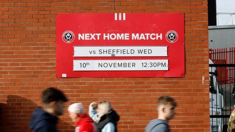 Football fans walk past a red sign at Bramall Lane reading: Next home match vs Sheffield Wed 10th November 12:30pm.