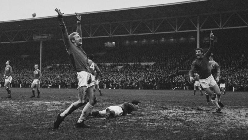 Denis Law, with short hair, holds his arms aloft after scoring for Manchester United. An opponent lies on the floor behind him and a teammate turns towards him, smiling and raising his arm