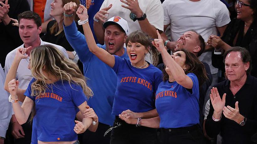 Taylor Swift and others are seen courtside cheering for the New York Knicks. She is wearing a blue T-shirt with orange lettering - the Knicks colors. 