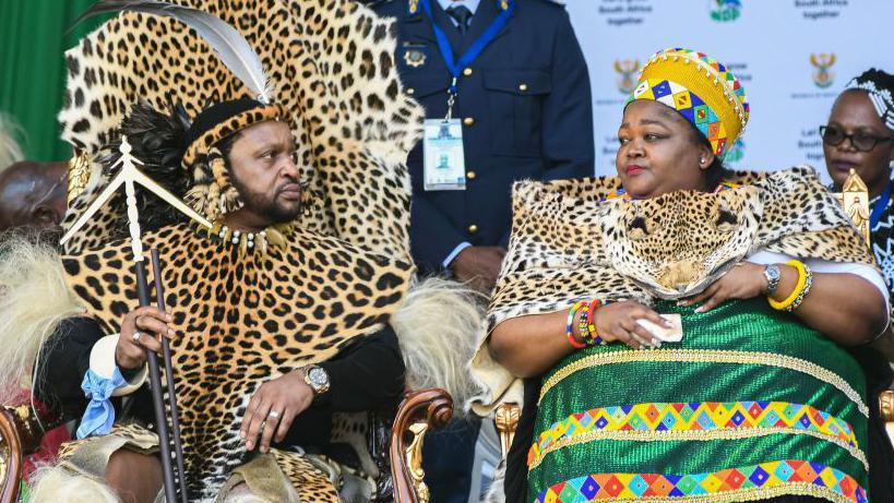 South Africa's Zulu king and his first wife are seen in colourful traditional attire during an official event. The king is wearing a leopard print wrap over his black long sleeved top while the queen is also wearing a similar wrap over her traditional green dress.