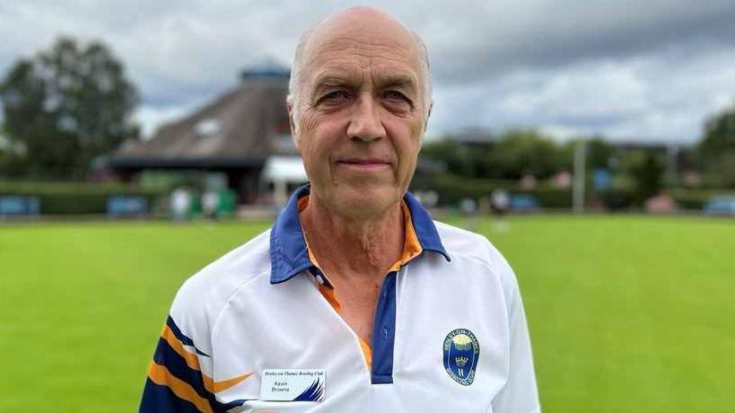 Kevin Browne smiling at the camera is wearing a white, blue and yellow bowling shirt with the bowling green in the background.