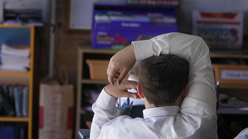 A child sat in a school classroom with their right arm draped over their head; the classroom has other students, books, shelves and boxes filled with teaching materials.