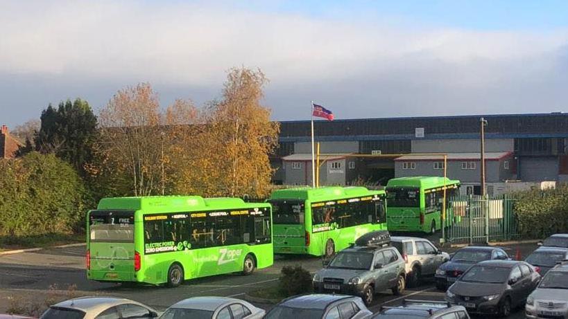 Three green buses can be seen parked up in a car park, with other vehicles nearby. There are trees and an industrial building in the background.