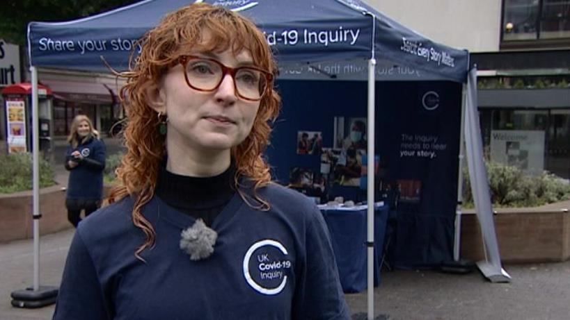 Kate Eisenstein - a woman with long curly red hair and glasses with red rims, wearing a a blue top that says "UK Covid-19 Inquiry". She is standing in front of a Covid-19 Inquiry stall. 