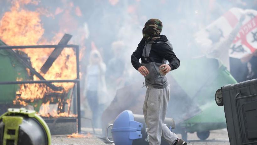 A man in a balaclava throws something with protestors and fire in the background. He is surrounded by rubble and turned-over bins