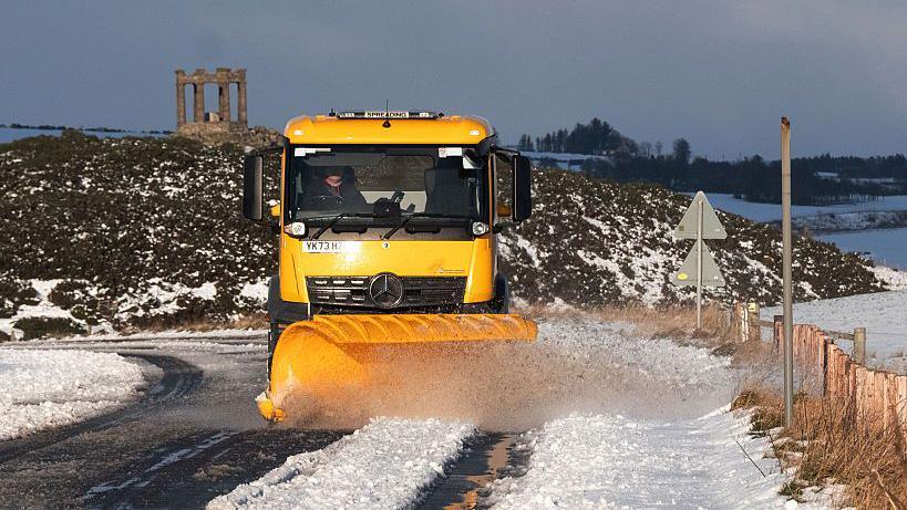 A snow plough - bright yellow in colour - clears snow from a rural road in the north east of Scotland