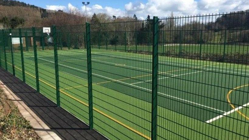 A CGI image showing a bright green all-weather sports pitch with floodlights and high fencing 