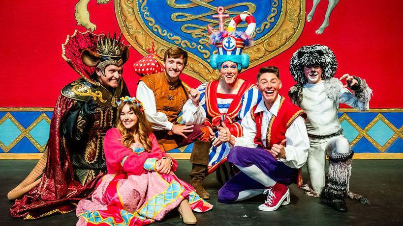 Six people sitting and kneeling on a stage dressed in colourful pantomime outfits. They are in front of the stage background, which is bright red, blue and gold at the bottom, with a large crest symbol.