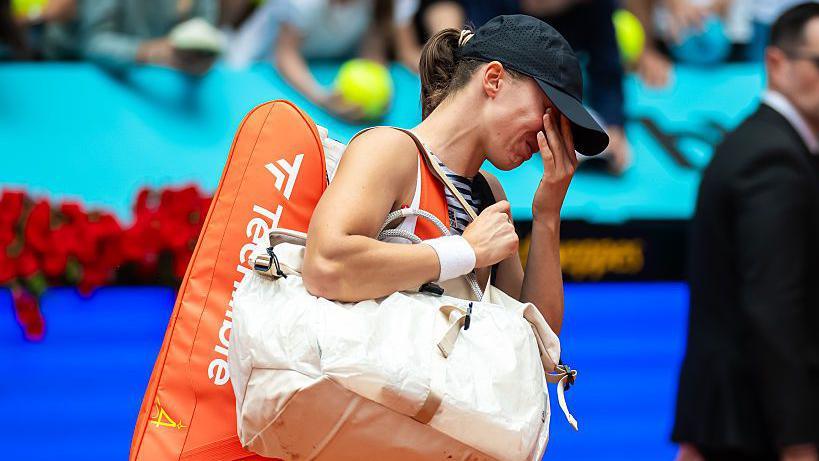 Iga Swiatek carries two bags and holds her hand to her face