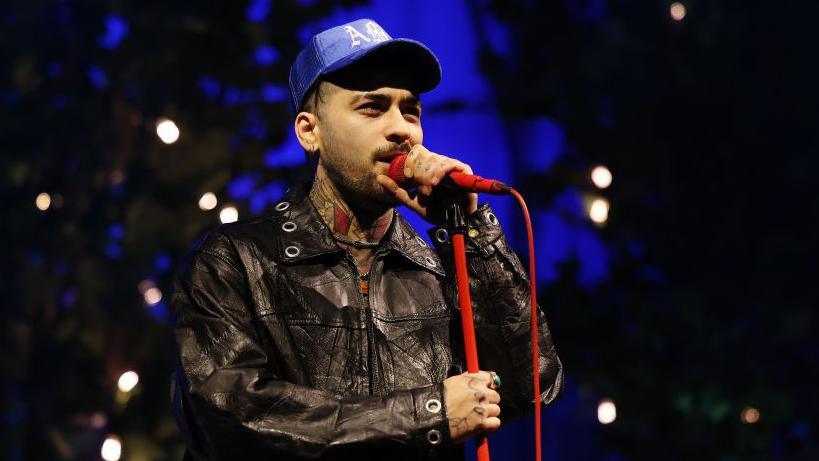 Zayn Malik wears a leather jacket and a blue hat. He is singing into a red microphone on a red microphone stand.