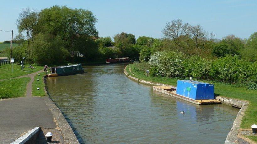 The Grand Union Canal at Three Locks, just outside Stoke Hammond. There are two long canal boats on the water on a sunny day. The canal is lined with trees either side and has a pathway to the left.