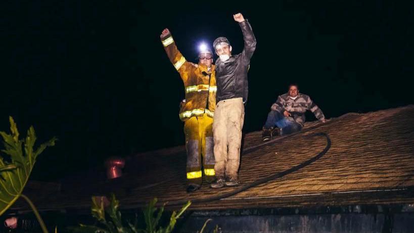 Two men stand on a roof with arms raised smiling at the camera. One is wearing a high-vis suit and the other a dark jacket stained with ash. Another man sits behind them wearing a lumberjack shirt