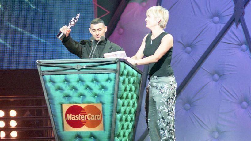 Two individuals on stage behind a podium with a MasterCard logo - one holding up an award.