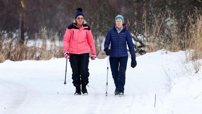 Two woman walking in the snow in Braemar. They are wrapped up in warm jackets, trousers, hats and gloves and wear sturdy boots. They are carrying walking poles. 