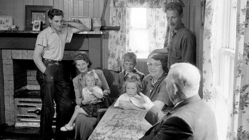 A family gathering pictured in the Western Isles in 1955. The family is gathered in a kitchen and the group comprises two men, two women and three children listening to an older man talk.