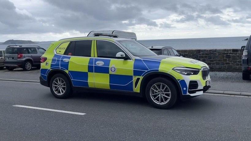 A Guernsey Police car, which appears to be a BMW, parked on a road next to a seawall on the island. The car has blue and yellow squares all over it. It is a cloudy day.