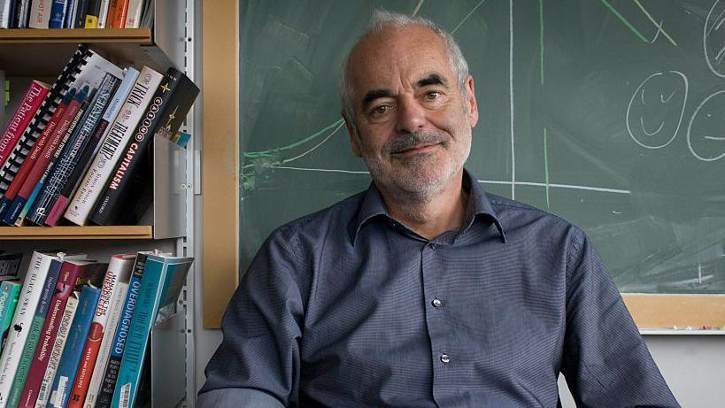 Prof Sir David Spiegelhalter wearing a blue shirt and sitting in front of a blackboard with books to his right.