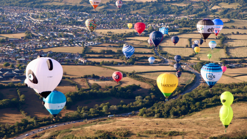 Many colourful balloons are seen flying over the outskirts Bristol and yellow/brown fields