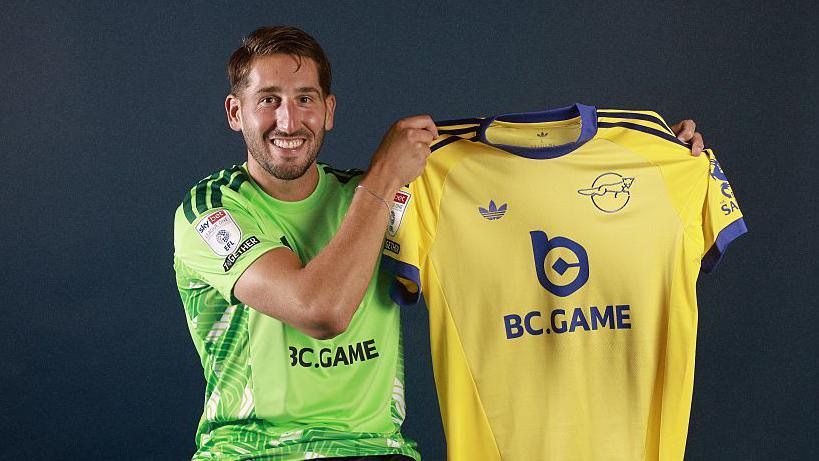 Franco Ravizzoli, sat down while wearing a green Leicester City goalkeeping jersey, holding a yellow Leicester City shirt up while smiling.