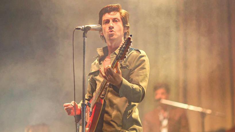 A man with brown wavy hair on stage, standing behind a microphone and holding a guitar. He is looking into the crowd.