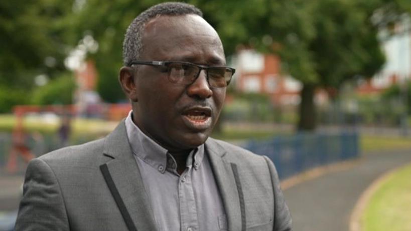 A man in a grey suit with glasses speaking on a street