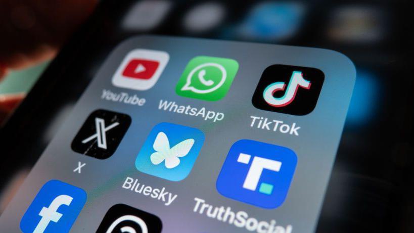 An iPhone screen is lit up and shows an open folder with various social media logo apps including YouTube, WhatsApp, TikTok, X, Bluesky, TruthSocial and Facebook