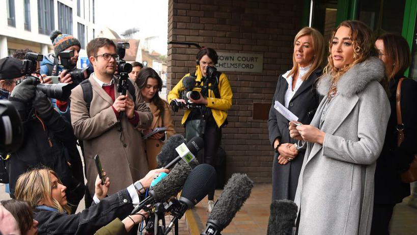 Georgia Harrison wearing a long grey coat and grey trousers, making a speech outside the glass doors of a court. Her lawyers are standing behind her and members of the press are standing in front of her holding out microphones.