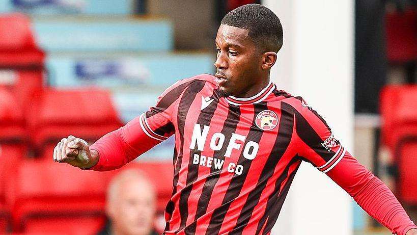 Walsall striker Daniel Kanu wearing home red and black stripes and concentrating on the ball just ahead of him out of shot