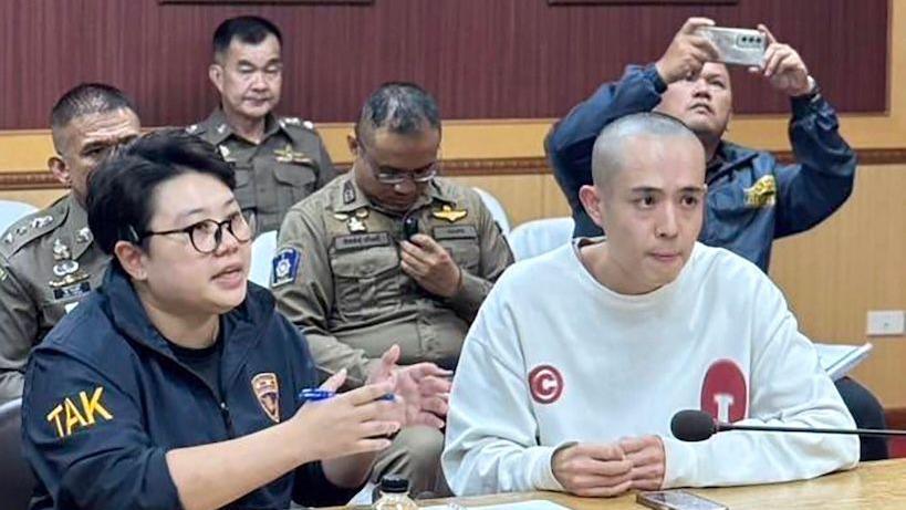 Wang Xing sitting on the right of a table in a white sweater and shaved head. Beside him is a person in navy uniform. Sitting behind are police officers in their uniforms.