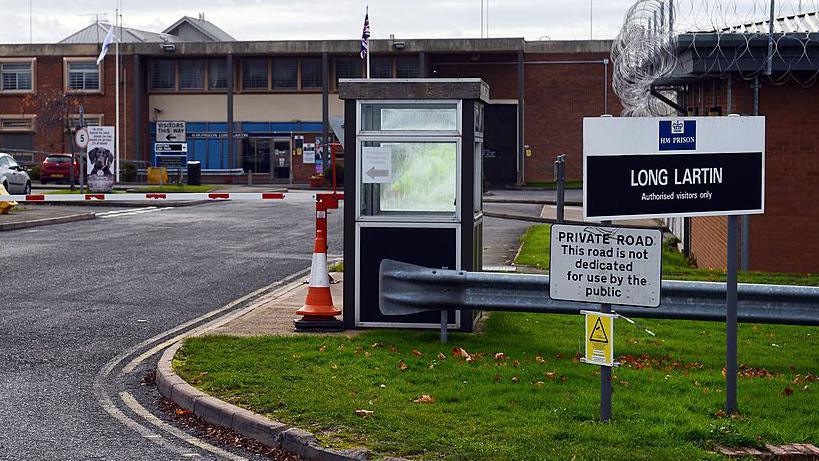 Exterior view of Long Lartin prison - sign with name and the road barrier into the prison car park