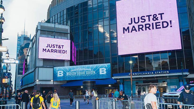A sign outside New York's Madison Square Gardens confirming Taylor and Travis got married. It says "Just T&T Married" on a large screen, with barriers in front and people hanging around outside