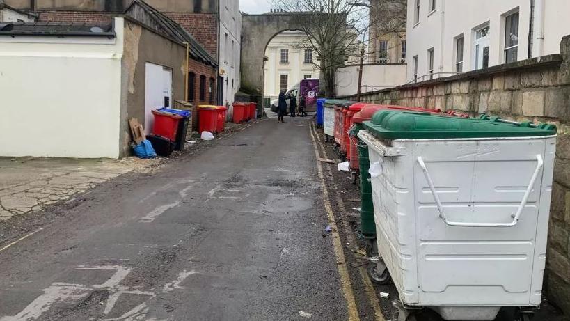 Street with lots of big bins and rubbish on the road near them. They are at the back of houses.