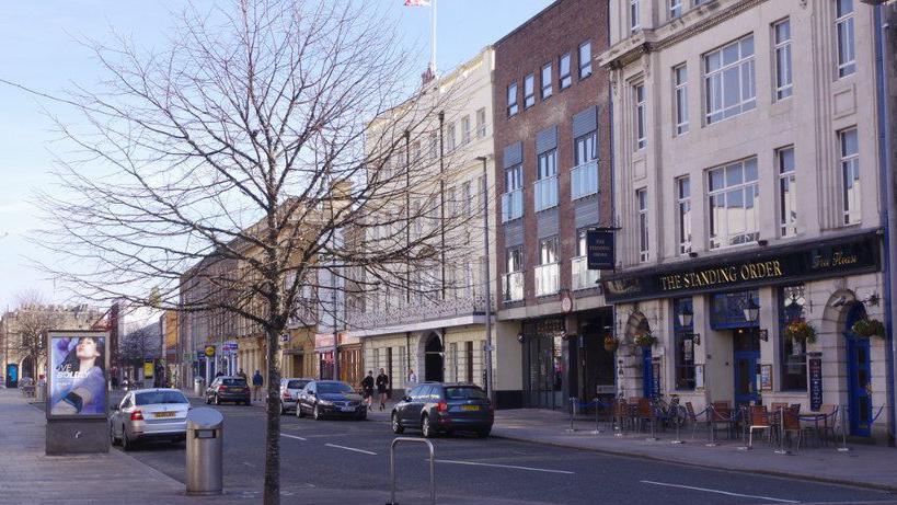 Wetherspoon pub The Standing Order as seen from the other side of the street. Chairs and tables are on the pavement outside the pub.