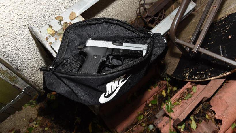 The 9mm handgun sitting inside an open black Nike bag, which has been hidden behind old furniture and debris in an alleyway. The picture is taken with a flash at night.