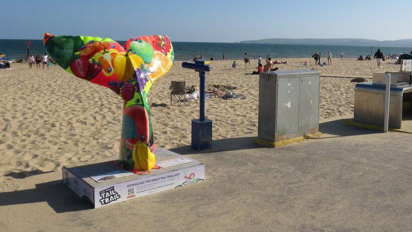 A mermaid tail sculpture on a plinth on the esplanade at Bournemouth. It is decorated with a montage of fruit and vegetables.