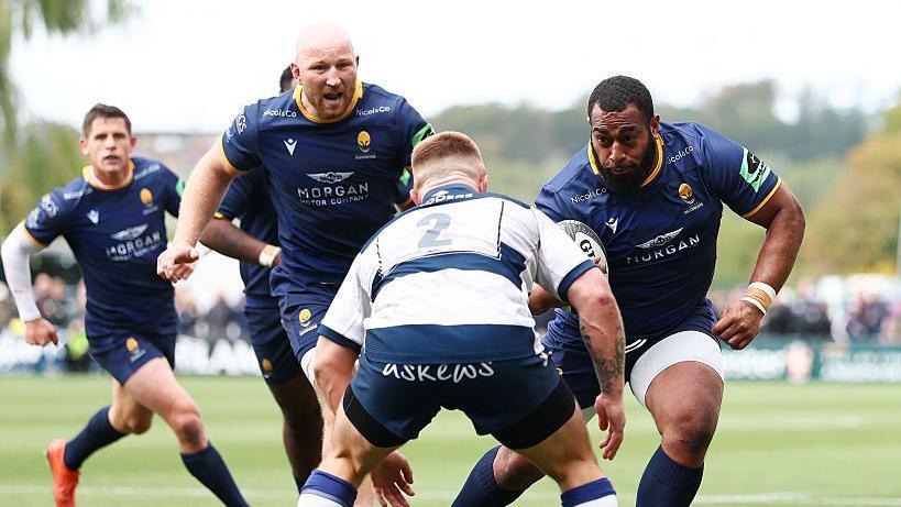 Two Worcester Warriors players in dark blue charge towards a lone Coventry defender in white.
