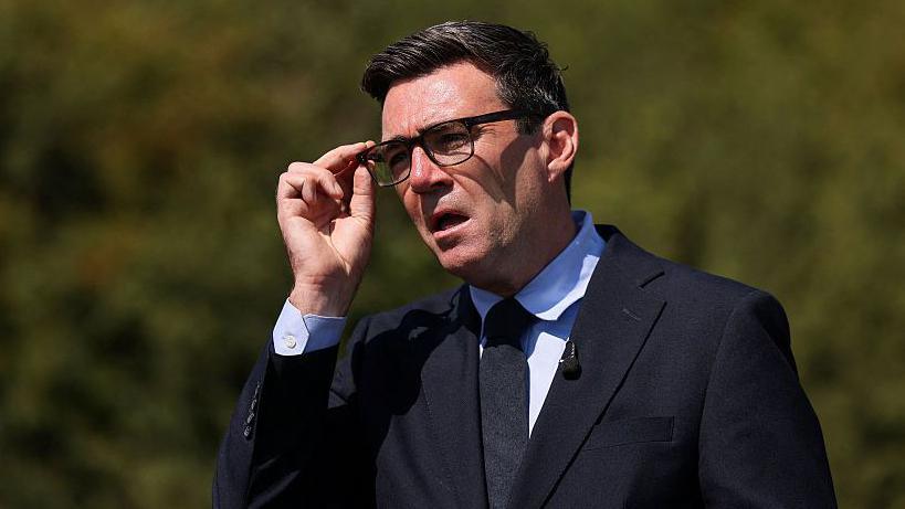 Andy Burnham wearing a suit. He is holding his glasses and speaking.