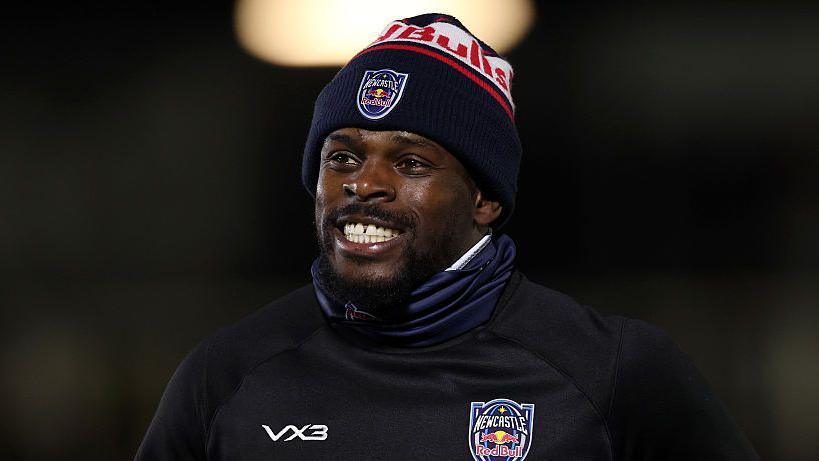 Christian Wade has a big smile as he warms up for a Newcastle match, wearing a woolly hat, snood and a big sweatshirt with the Newcastle Red Bulls badge on it 