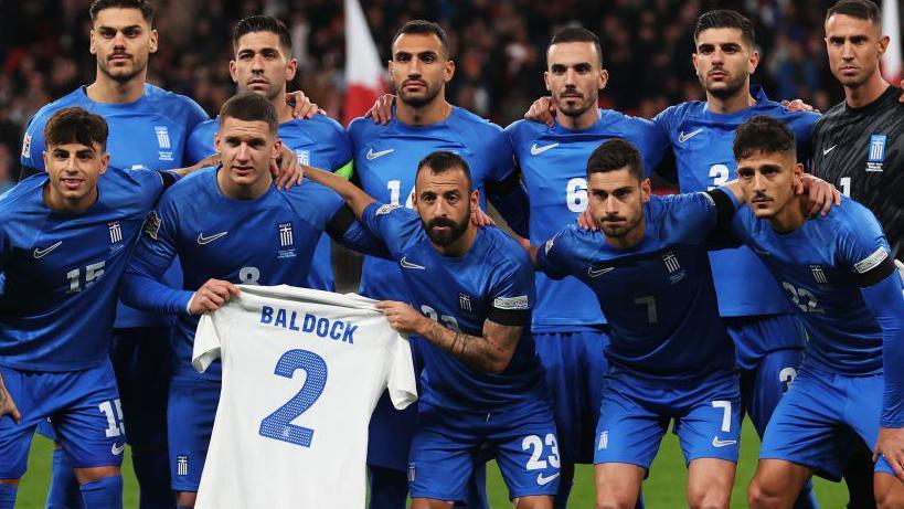 Greece players line up behind a shirt bearing George Baldock's name before the match against England