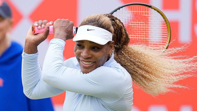 Serena Williams, wearing white and smiling, plays a backhand