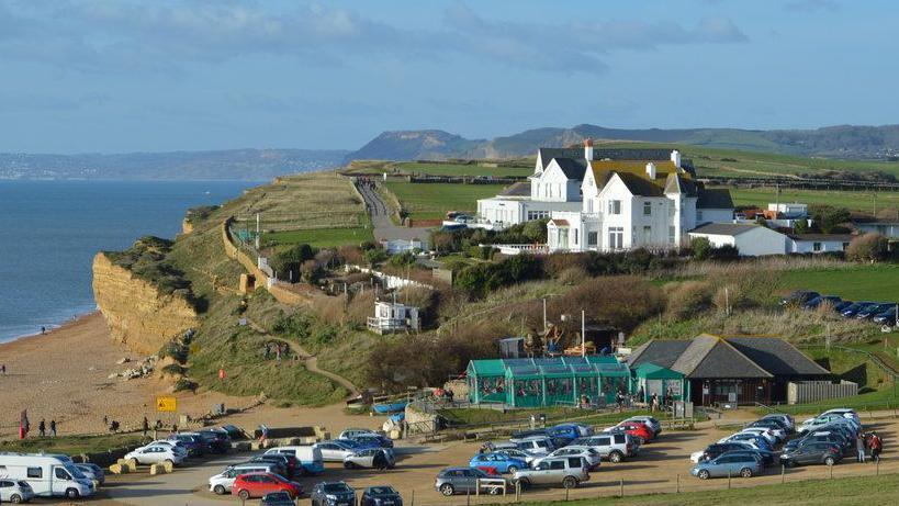 The view across Hive Beach car park towards Hive Beach Cafe, with the white Victorian house and the Seaside Boarding House guesthouse on the low cliffs beyond. The car park in the foreground is full of cars. To the left is the beach and the sandstone cliffs that gradually rise up into the distance. The beach cafe sits below the Victorian buildings on the edge of the car park and is a low single-storey building with a green-covered outdoor seating area.