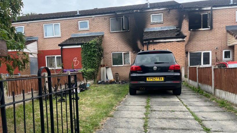 Two terraced houses with visible smoke damage on the brickwork and windows. Some charred and blackened furniture and goods are visible on the grass of the front lawn and a black Volkswagen is parked on the drive. 