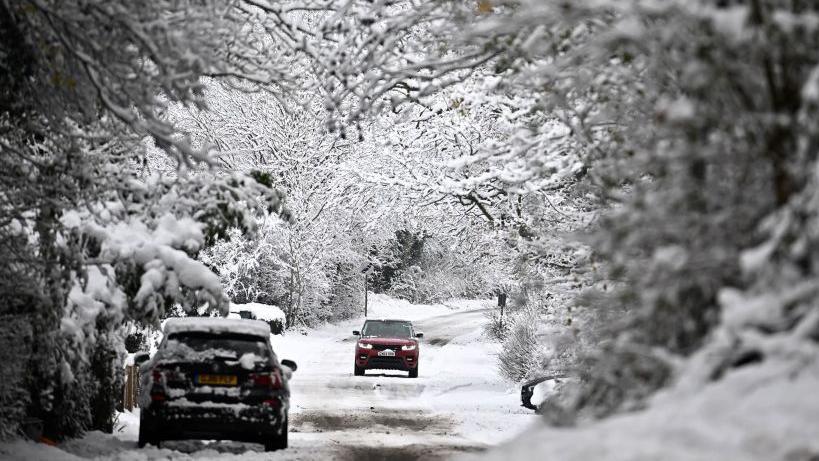 A car drives down a snowy country road which is framed by trees. Another car is parked at the side of the road.