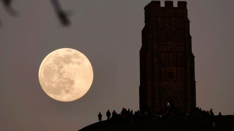 Një hënë e plotë lind në të majtë të një strukture të ngjashme me kullën në një kodër. Silueta njerëzish që qëndrojnë në fund të kullës.