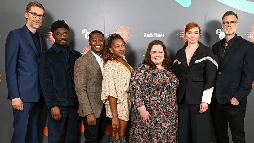 Stars of the Outlaws cast stand side-on to the camera in formal wear.