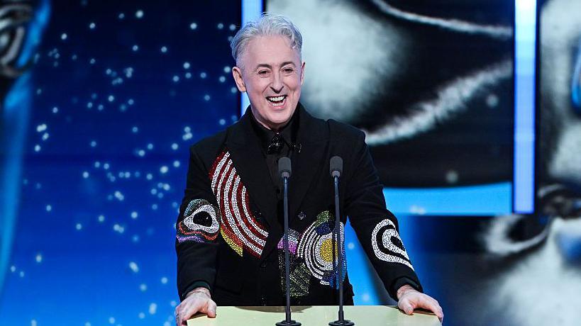 Alan Cumming flet në dy mikrofonë që ndodhen në një podium, të cilët i mban me të dyja duart.