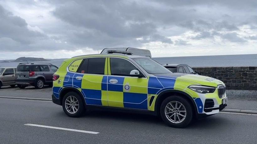 A police car is driving along a coastal road with cars parked along the sea wall. The sea can be seen beyond the wall and part of the mainland in the distance.