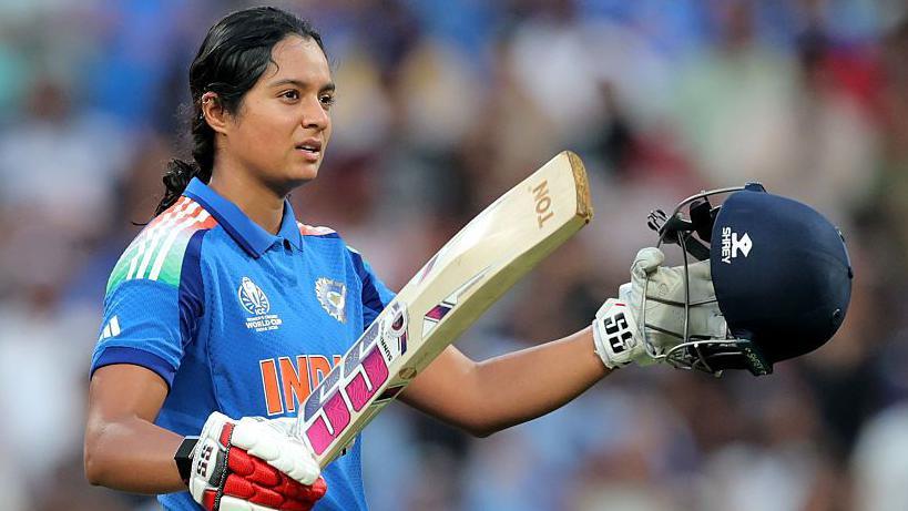 Pratika Rawal celebrates scoring a century in a One-Day international for India. She is holding up her helmet in her left hand and bat in her right. She is wearing a blue India shirt