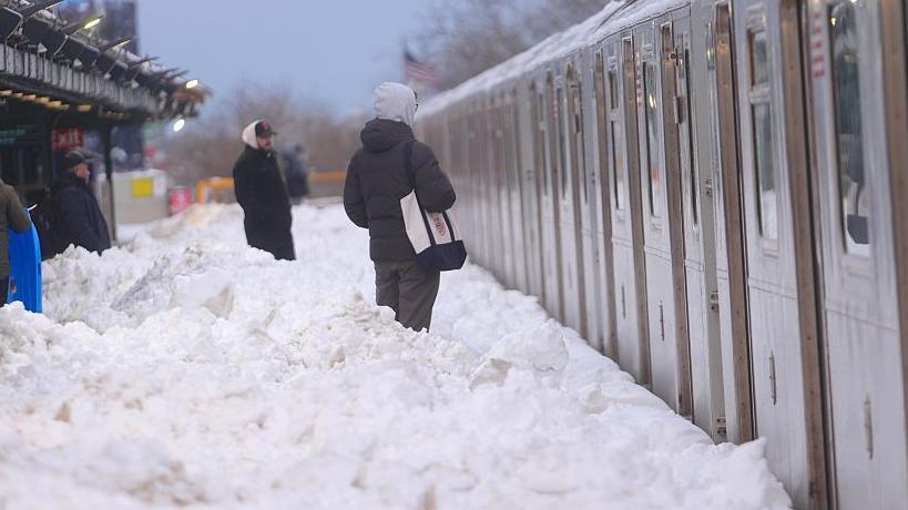 Three people can be seen standing up to their knees in snow on a train platform. Train carriages are to the right of the photo.